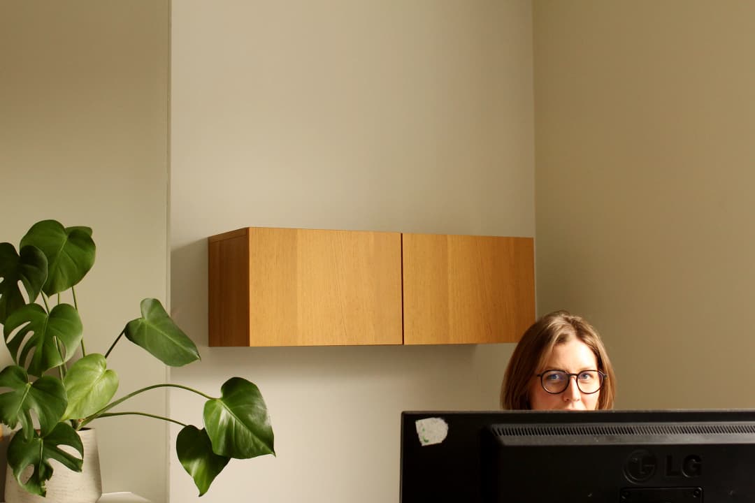 Project manager Katy looking at camera over monitor, beside houseplant